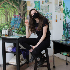 Our Member of the Month, Jocelyn sitting on a stool in her studio They are holding a peace sign.
